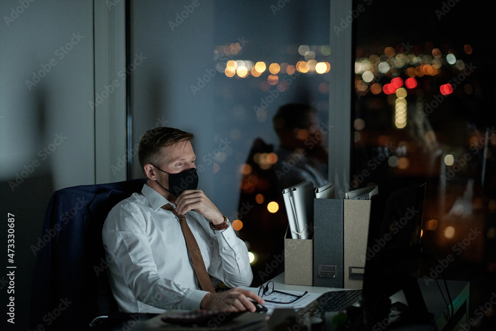 Poster Young white collar worker in protective mask looking at computer screen while sitting by desk and working late