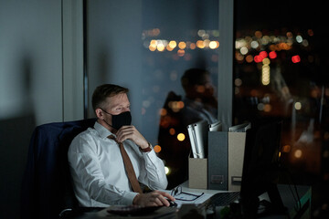 Young white collar worker in protective mask looking at computer screen while sitting by desk and...
