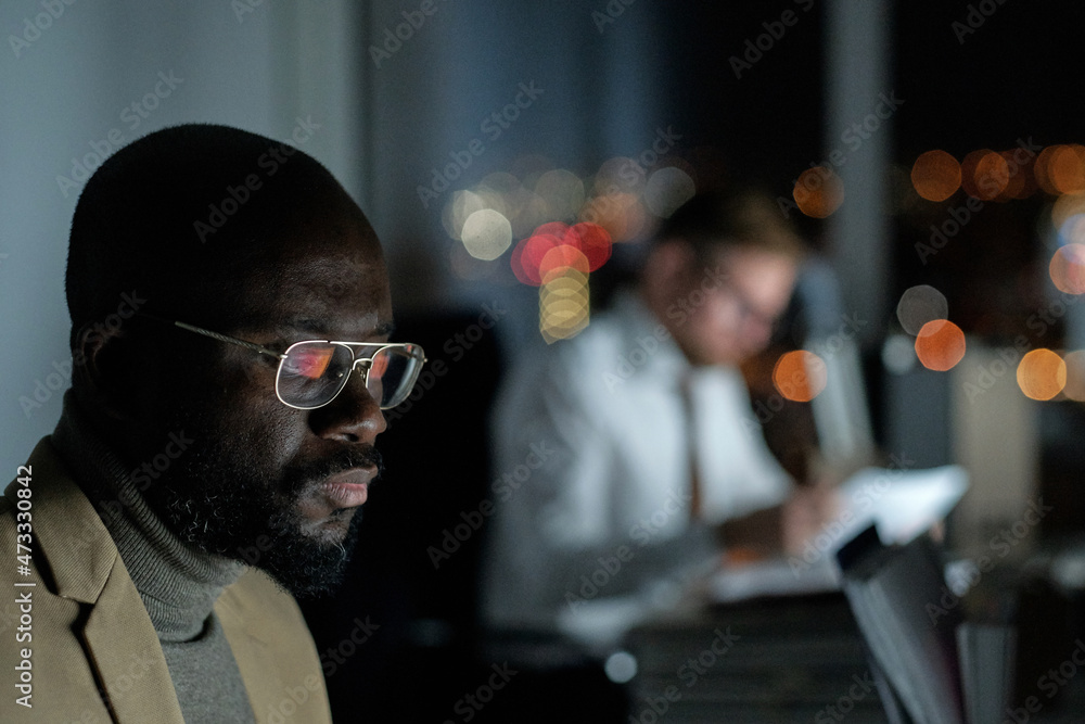 Poster young serious businessman in eyeglasses looking at computer screen while working late in the evening