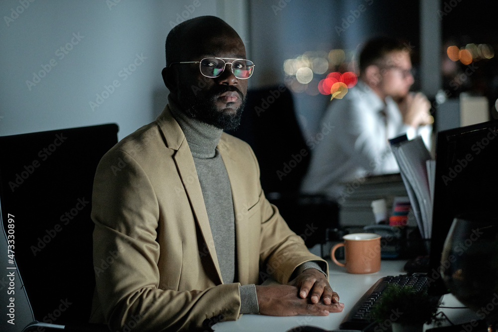 Poster african businessman in formalwear looking at you while sitting in front of computer monitor by workp