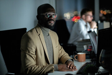 African businessman in formalwear looking at you while sitting in front of computer monitor by workplace against colleague