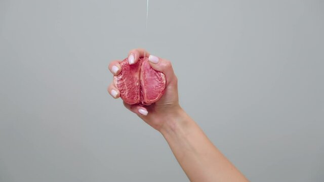A woman's hand clutches half a grapefruit in her fist. Liquid is poured on the fruit. Close up. Gray background. The concept of female sexuality.