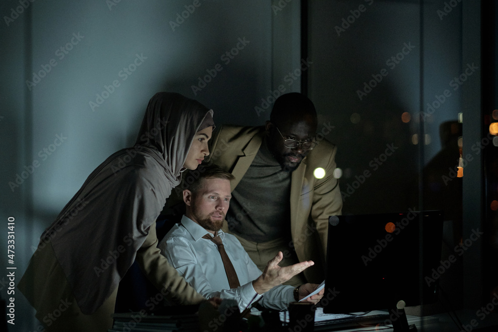 Sticker Three young intercultural colleagues looking at computer monitor during presentation at night business meeting