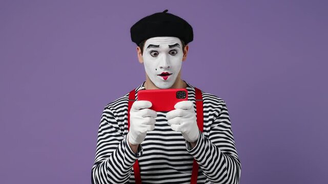 Gambling young mime man with white face mask wears striped shirt beret play racing on mobile cell phone hold gadget for pc video games isolated on plain pastel light violet background studio portrait