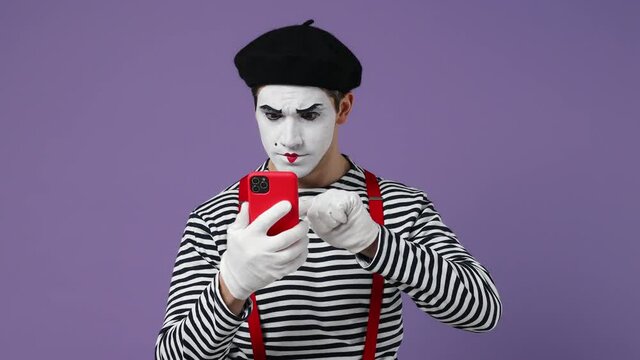 Dreamful pensive young mime man with white face mask wears striped shirt berethold using mobile cell phone typing browsing send sms isolated on plain pastel light violet background studio portrait