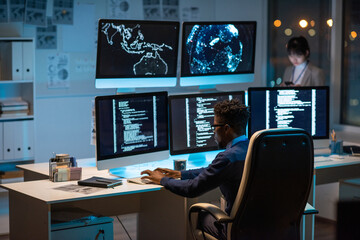Young contemporary cyber security manager typing while sitting by desk in front of computer monitors