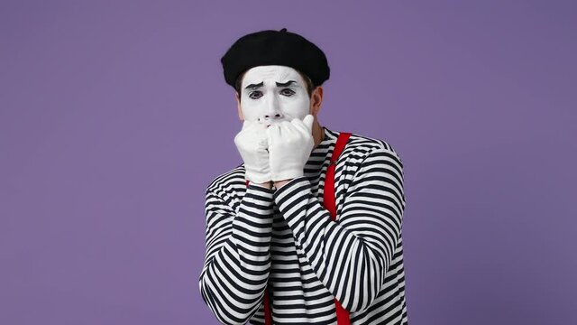 Scared shocked young mime man with white face mask wear striped shirt beret look camera cover hide face with hands peep through fingers isolated on plain pastel light violet background studio portrait