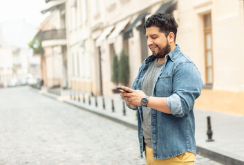 Young cheerful hispanic man in jeans jacket using phone on european street
