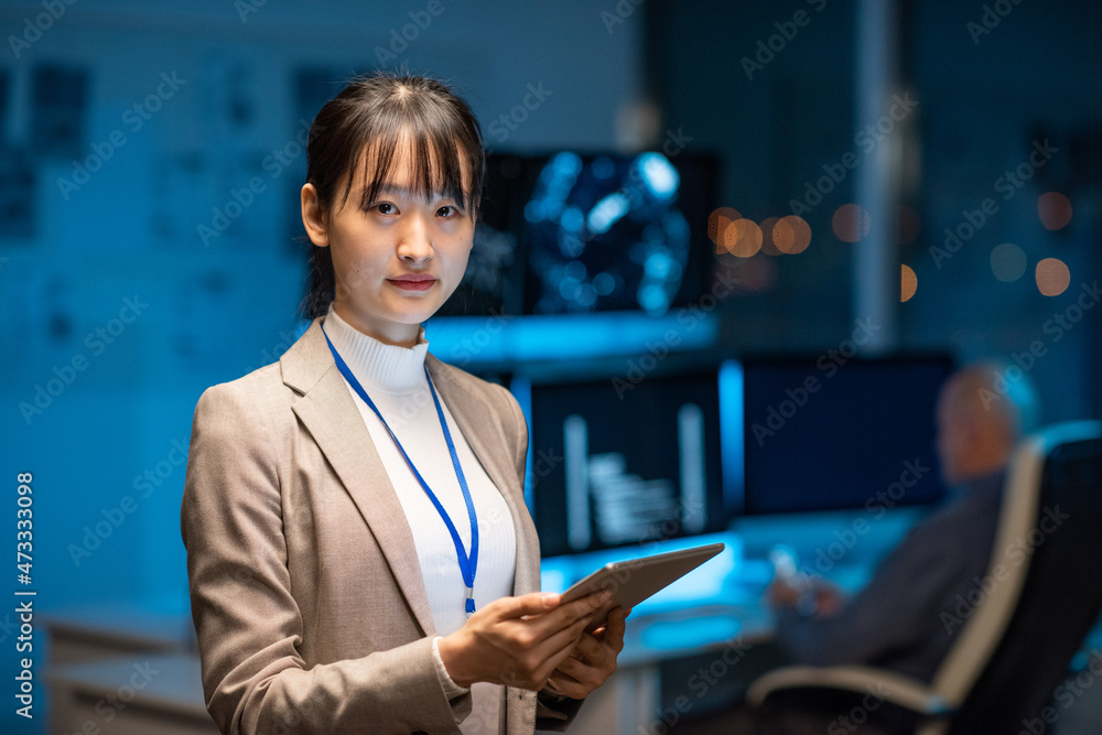Poster Young serious female programmer or developer in formalwear holding tablet while networking in office environment