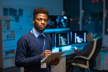 Young serious African programmer with tablet standing in front of camera against colleague by...