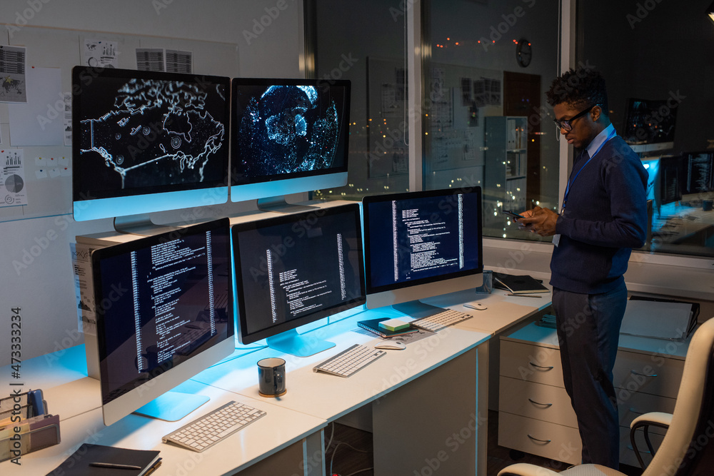 Poster Young African programmer texting in smartphone while standing by workplace against large office window at night