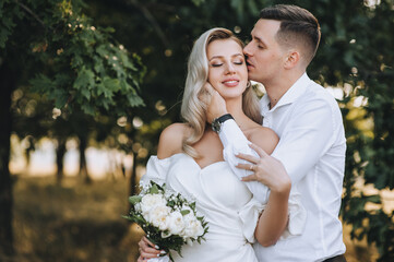 Smiling groom and a beautiful blonde bride with curly hair in a white dress tenderly hug in nature...