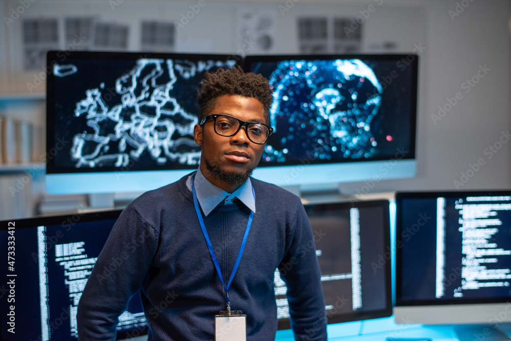 Poster Contemporary young programmer standing by workplace against group of computer monitors with coded data on screens