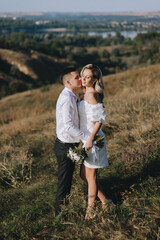 A loving groom and a beautiful blonde bride in a white dress tenderly hug in nature on the background of hills and cliffs in autumn in sunny weather. Wedding photography.