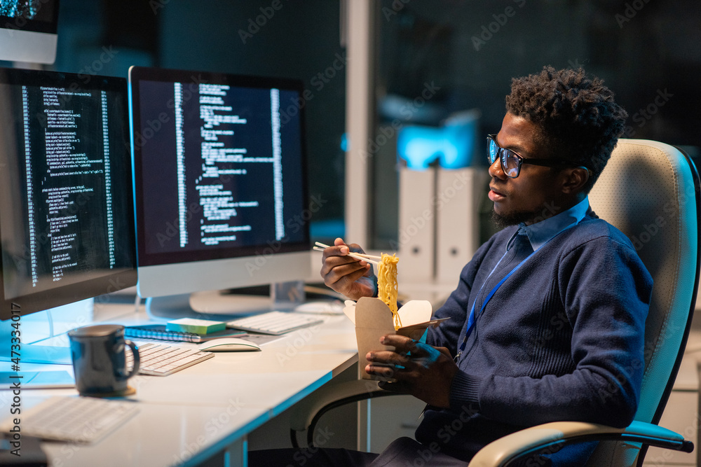 Poster Young hungry programmer eating chinese wok in front of computer screen with coded data while working in office at night