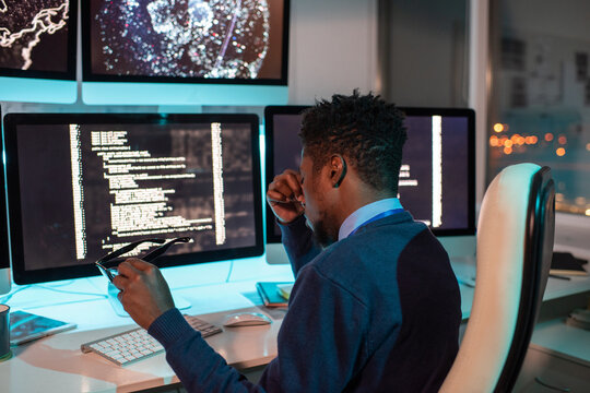 Young tired African businessman sitting in front of computer monitor with coded data on screen while working overtime