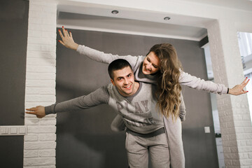 Happy young couple man and woman enjoy leisure time together. The girl and her boyfriend are dressed in home clothes. A cheerful girl climbed onto the back of her boyfriend and spread her arms