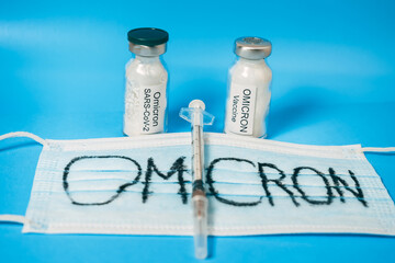 A medical mask labeled Omicron, a syringe, and two vials of vaccine. The vaccine is for a new strain of covid-19 coronovirus. Blue background.