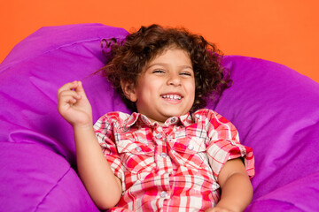 Photo of funny pretty school girl dressed checkered clothes lying bean bag smiling isolated orange color background