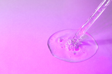 Facial serum in glass pipette in pink neon light closeup. Liquid gel pouring from transparent dropper with bubbles. Front view. Trendy cosmetic product with hyaluronic acid macro. Copy space.