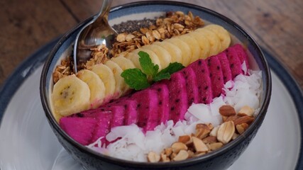 Lifting metal spoon from berry smoothie bowl with granola, banana, pitaya, coconut, nuts and seeds; superfood healthy vegan breakfast 