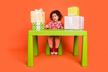Photo of moody upset schoolgirl wear plaid outfit sitting green table dislike gifts boxes isolated orange color background