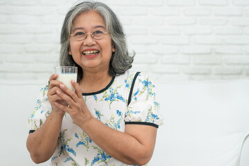 Asian Senior woman holding glass milk while relaxing on a sofa living room for retirement wellness....
