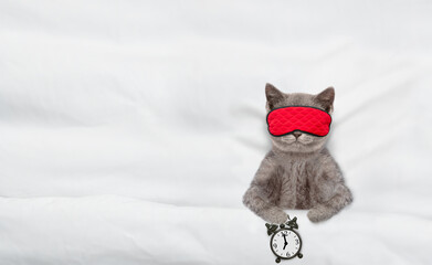 Cute kitten wearing mask for sleep shows alarm clock on a bed at home. Top down view. Empty space...
