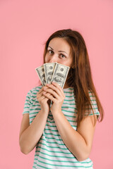 A beautiful brunette woman in casual clothes won a small amount of money in a lottery. Portrait of a girl with dollar bills covering her face. Vertical photo on pink background