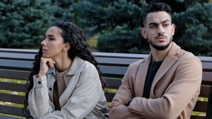 Offended young hispanic couple looking different direction sitting on bench in autumn park annoyed...