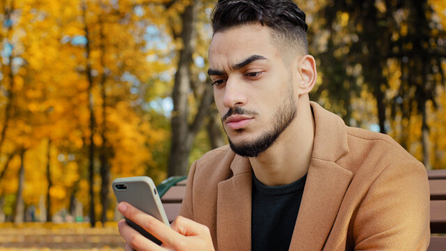 Portrait Young Serious Guy Use App Hispanic Man Looking At Phone Screen Browses News On Telephone Concentrated Male Sits On Bench In Autumn Park Read Using Device Businessman Holds Smartphone Outdoors