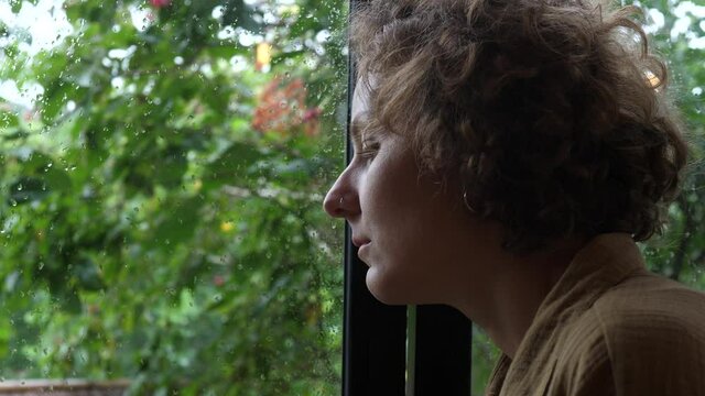 Portrait of lonely female thoughtfully looking through the window on a rainy day; feeling sadness, grief, and boredom