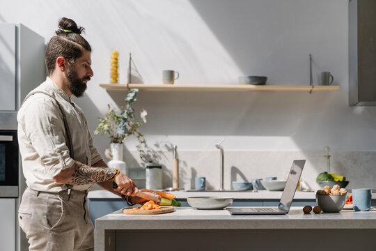 Bearded Man Cutting Vegetable While Looking At Laptop In Kitchen