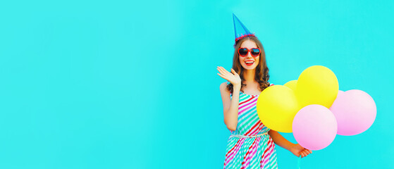 Portrait of beautiful happy smiling young woman with bunch of balloons in birthday cap wearing a colorful dress on blue background