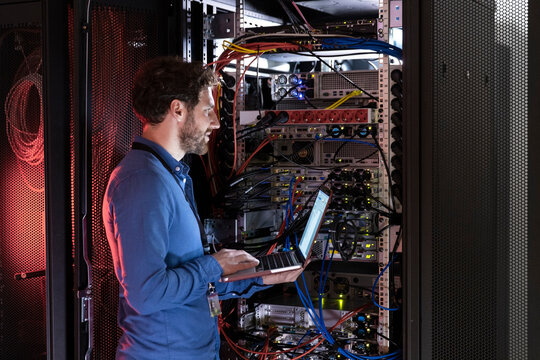 Male IT expertise with laptop looking away while standing at rack in data center