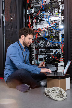 Smiling male IT specialist holding patch cord cable while using laptop in data center