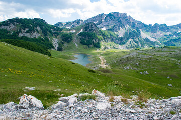 Mountain range and small glacial lake with background of green hills and cloudy blue sky. Durmitor National Park. Montenegro.