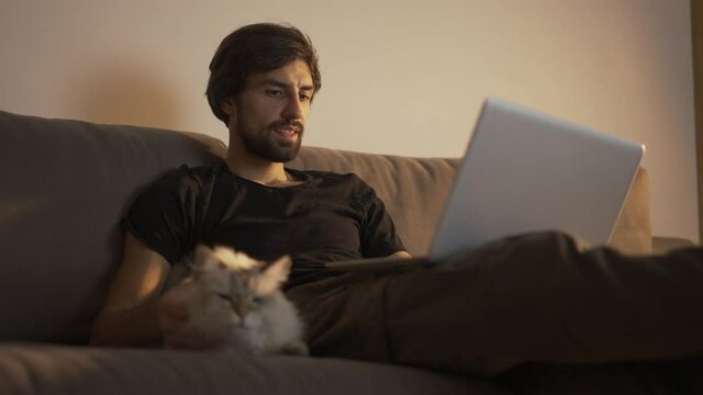 Man working on a laptop at home while sitting on the couch and petting a cat