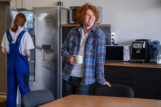 Smiling apprentice having coffee while female colleague searching in refrigerator at cafeteria