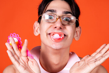 Portrait of a funny young man with a pacifier in his mouth and a rattle in his hands on an orange...