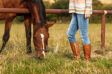 Woman with riding boots standing on pasture with her grazing horse