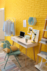 Fashion designer's workplace with wooden furniture and laptop near yellow brick wall. Stylish interior