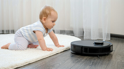 Cute baby boy looking with interest on robot vacuum cleaner on floor at living room. Concept of...