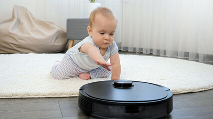 Cute baby boy crawling on floor and looking on robot vacuum cleaner. Concept of hygiene, household...