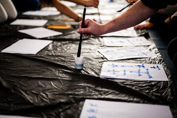 Unrecognizable man's hand dipping  paint brush in ink bottle on Japanese calligraphy workshop class, handwriting letters with group of classmates