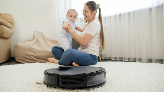 Robot Vacuum Cleaner Helping Woman Doing Chores While She Spend Time With Baby Son. Concept Of Hygiene, Household Gadgets And Robots At Modern Life.