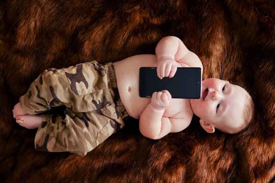 Funny happy baby boy with a phone in his hands on a fur carpet. Smiling child holds a smartphone, top view