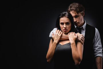 man in vest and shirt hugging and choking sexy girlfriend in silk slip dress isolated on black.