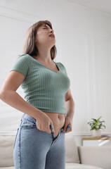 Young woman struggling to put on tight jeans at home