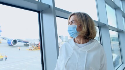 adult Female wearing protective mask is standing at the window in the airport terminal awaiting the departure of a flight due to travel restrictions due to the coronavirus pandemic, a senior aged 50